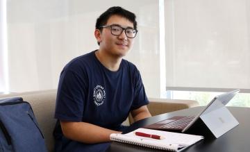 身着网络安全夏令营t恤的希望学者Tytain Sun坐在桌前，桌上放着电脑、笔记本和笔